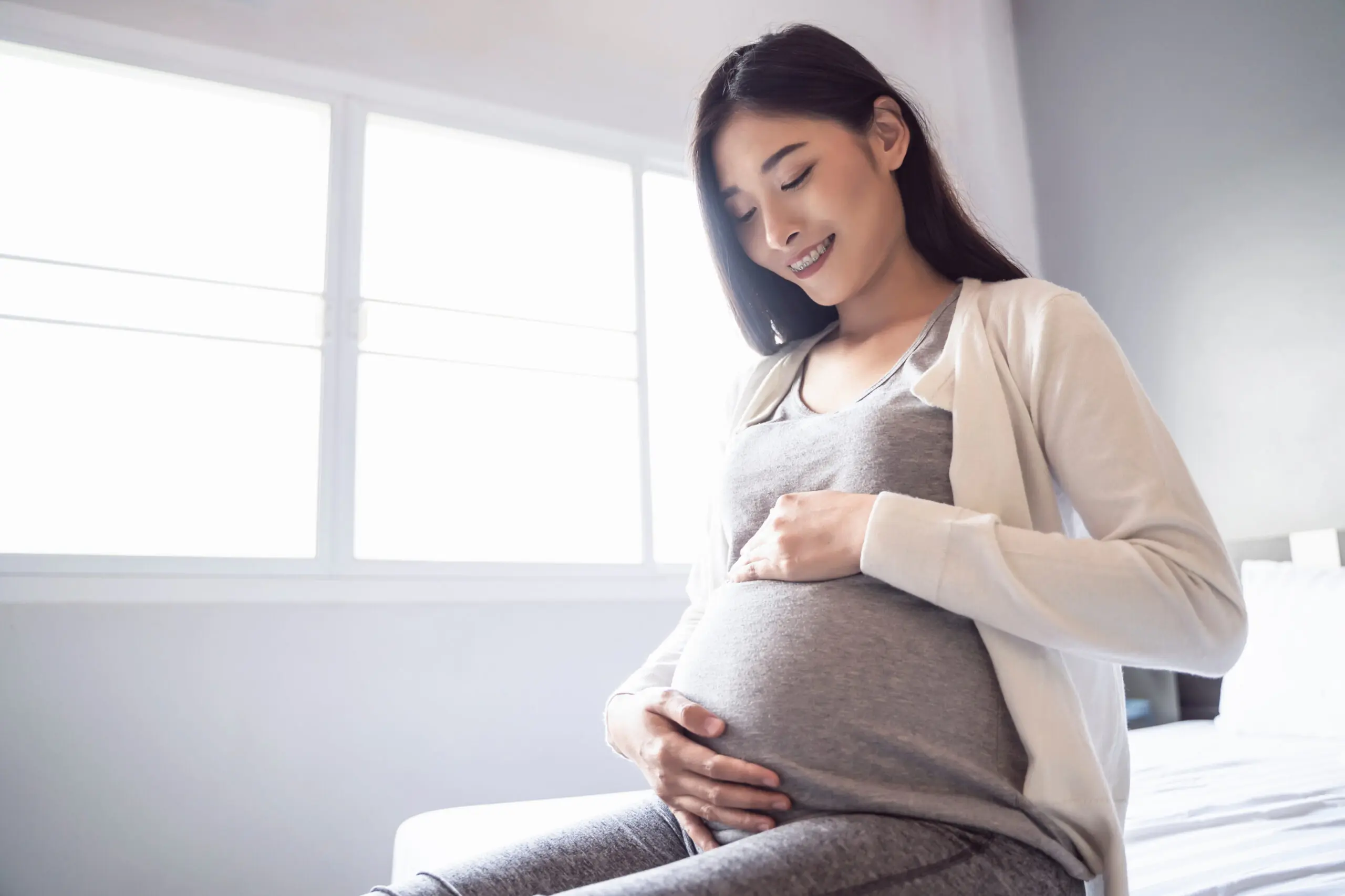 Asian woman sitting on a bed, looking down and smiling at her pregnant belly that she is cradling.