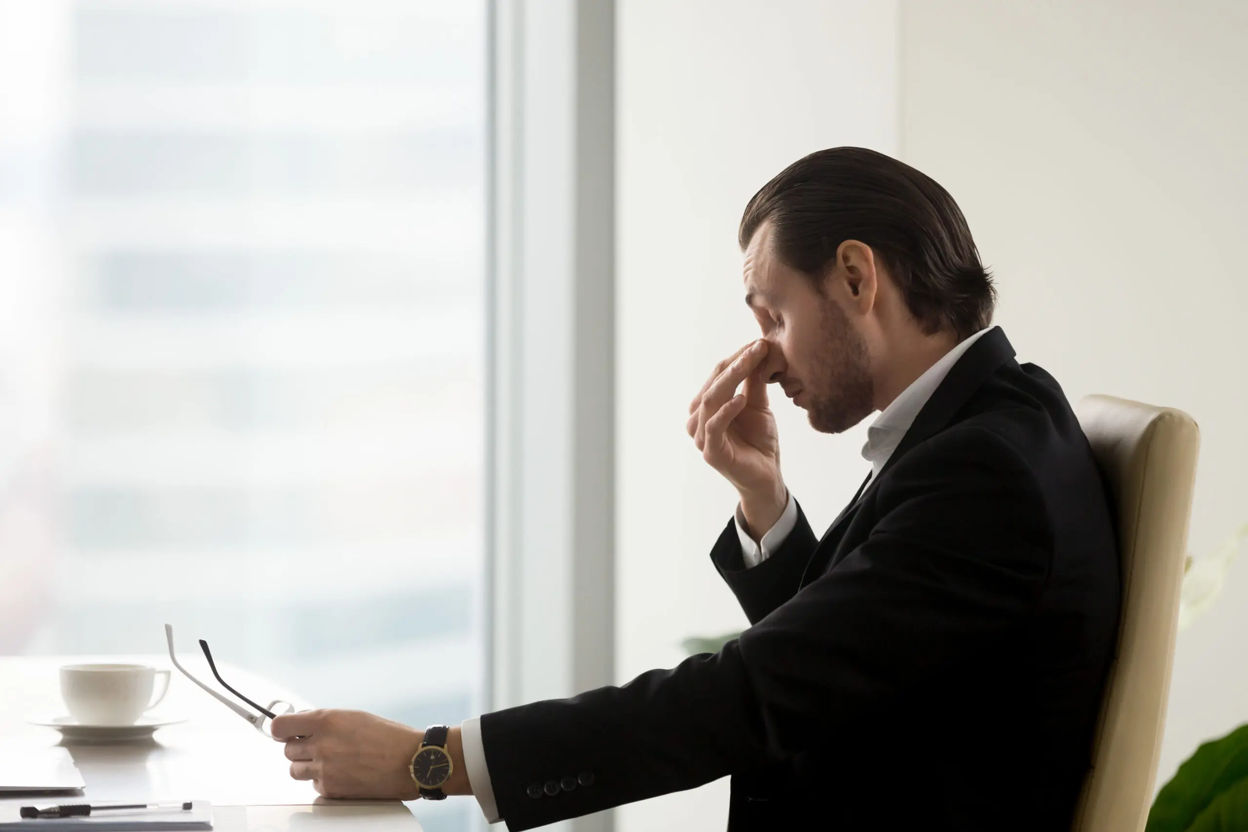 Professional man who feels exhaustion and burnout is holding the bridge of his nose between his fingers and has his eyes closed.